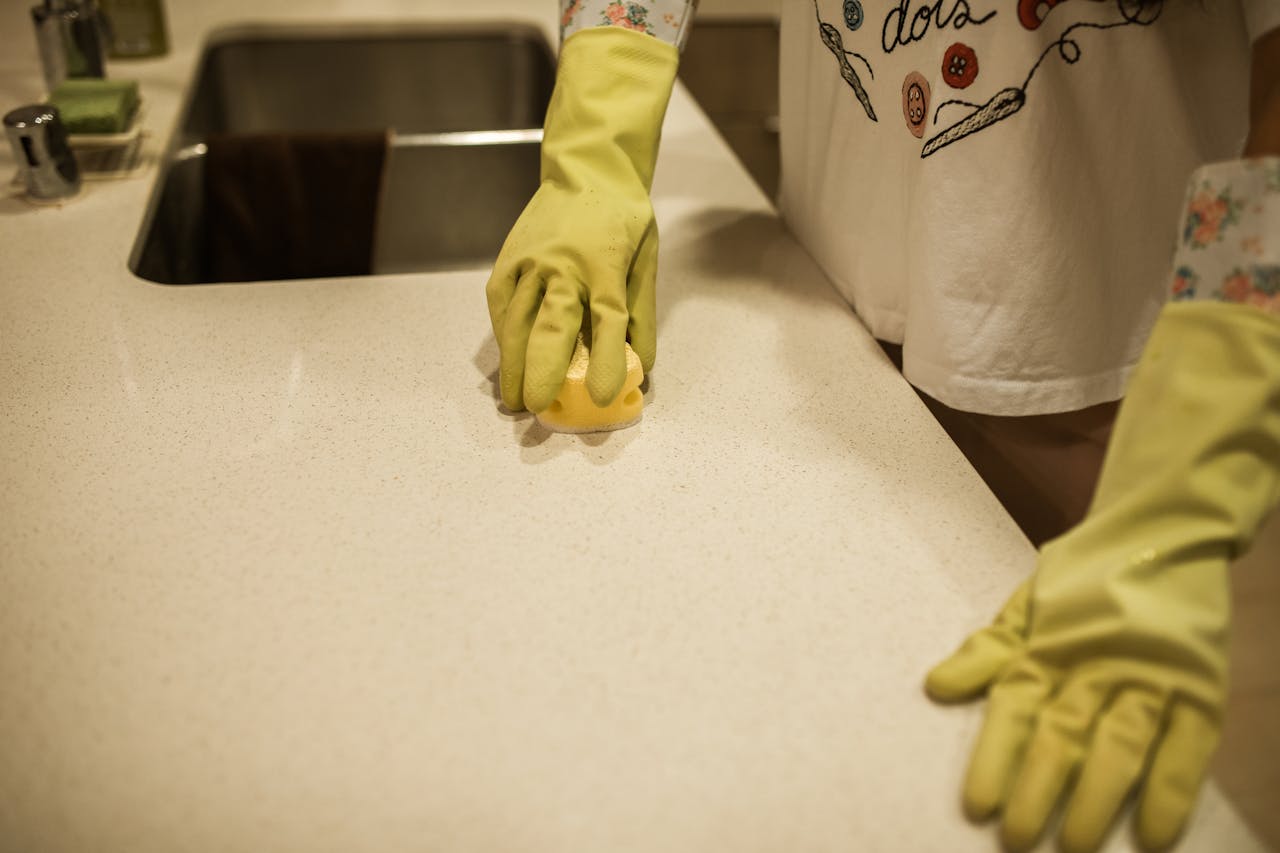 expertise-01 Woman using yellow rubber gloves to clean a kitchen counter with a sponge.